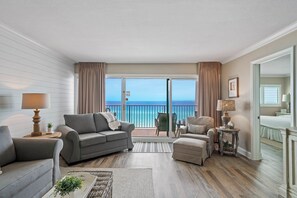 A living room with a sofa, armchairs, and a small table with a lamp. There is a view of the ocean through large windows.