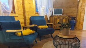 A cozy room with wooden walls, two blue armchairs with yellow cushions, a small round table with a plant, and a television on a wooden cabinet. - Chalet Cosy Edenna (Villefranche-sur-Cher)