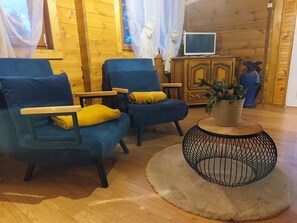 Een gezellige kamer met houten wanden, twee blauwe fauteuils met gele kussens, een klein rond tafeltje met een plant en een televisie op een houten kast.