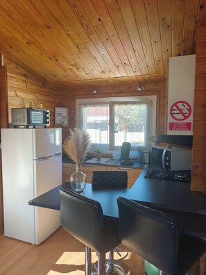 A wooden cabin interior with a kitchenette, a dining table with chairs, a refrigerator, a microwave, and a no-smoking sign. - Chalet Cosy Edenna (Villefranche-sur-Cher)