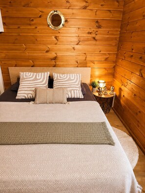 A wooden-paneled room with a bed, pillows, a round mirror, and a small table with decorative items. - Chalet Cosy Edenna (Villefranche-sur-Cher)