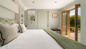 A bedroom with a large bed, a wooden side table, a mirror, and a door leading to a garden. - Dog friendly, two bedroom holiday cottage in heart of Cotswolds (Painswick)