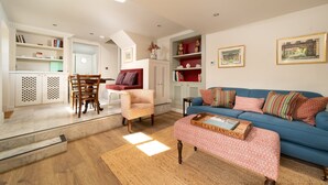 A living room with a blue sofa, patterned ottoman, wooden coffee table, and a dining area with a wooden table and chairs. - Dog friendly, two bedroom holiday cottage in heart of Cotswolds (Painswick)