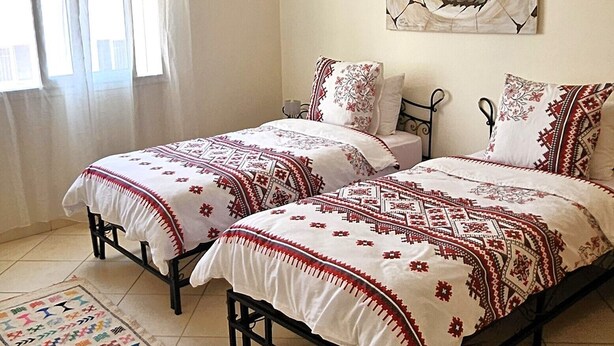 Quarto com duas camas, uma janela grande com cortinas leves e uma pintura de praia na parede, além de uma luminária de tecido.