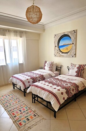 A bedroom with two beds, a large window with sheer curtains, a beach painting on the wall, and a woven light fixture. - Downtown apartment (Agadir)