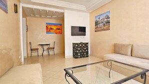 Uma sala de estar com uma mesa de centro de vidro, sofá bege e uma TV montada na parede. Há duas pinturas nas paredes e uma pequena mesa com cadeiras ao fundo.