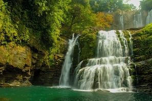 Uma cascata em vários níveis deságua em uma piscina turquesa cristalina, rodeada por vegetação exuberante.