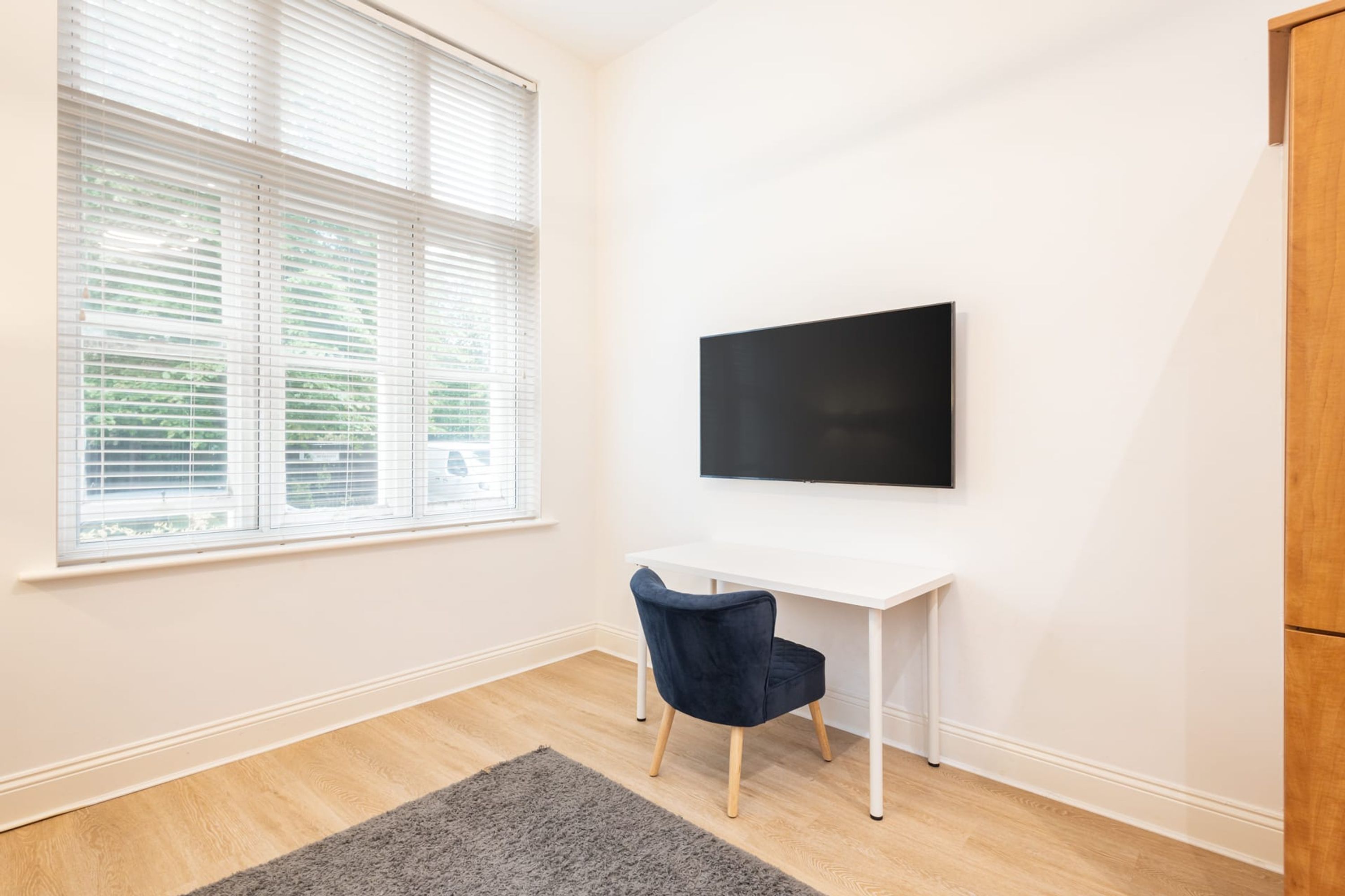 A room with a white desk, a dark blue chair, a black TV on the wall, and a window with blinds.