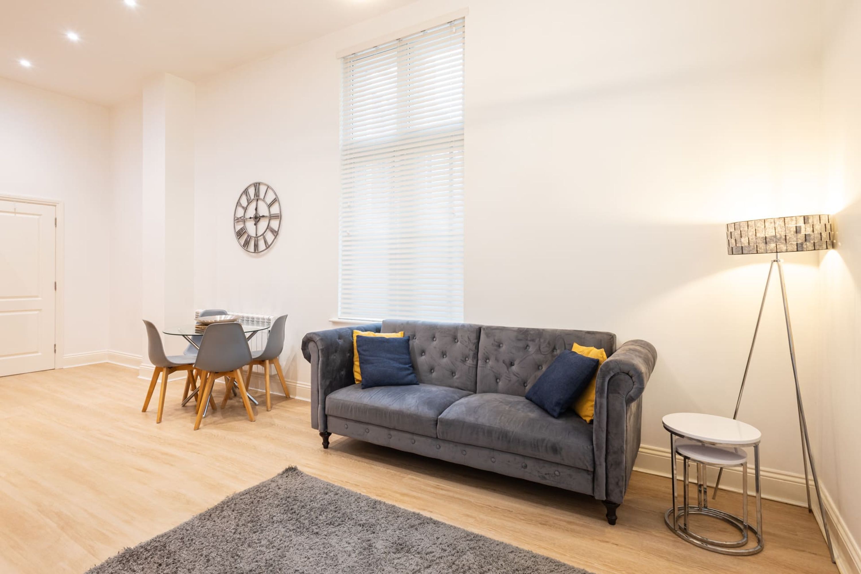 Ein Wohnzimmer mit grauem Sofa, einem kleinen runden Tisch, einem Esstisch mit Stühlen, einer Wanduhr und einer Stehlampe.