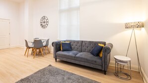 A living room with a grey sofa, a small round table, a dining table with chairs, a clock on the wall, and a floor lamp. - Host Stay The Cathedrals (Durham)