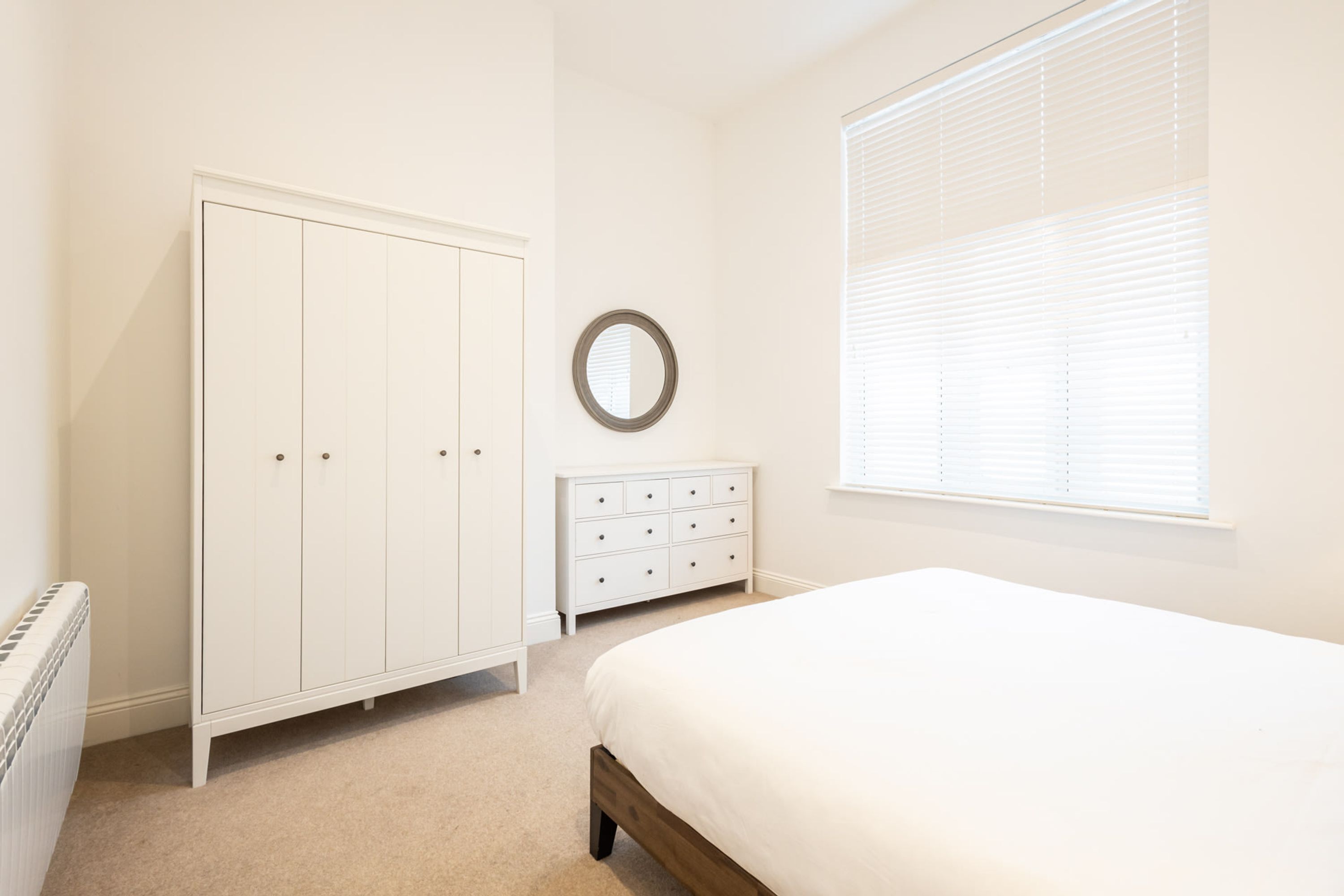 Een slaapkamer met een bed, een dressoir, een kledingkast en een raam met jaloezieën.