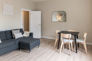 A modern living room with a grey sectional sofa, a small ottoman, a dining table with four chairs, and a white door.