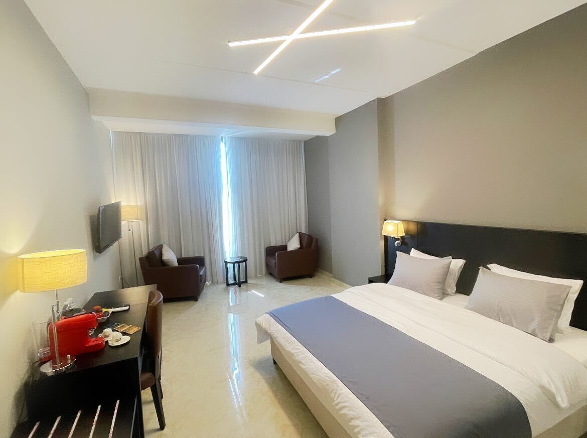 A modern hotel room with a large bed, a desk with a lamp, two armchairs, and a television.