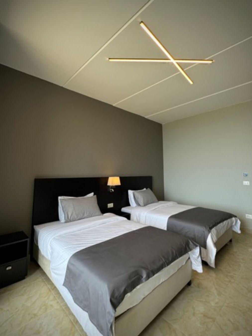 Una habitación de hotel moderna con dos camas, un cabecero oscuro, una mesita de noche con lámpara y un aplique de techo con diseño geométrico.