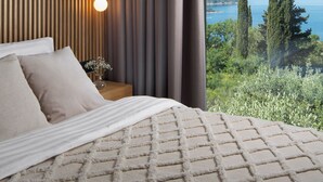 A bedroom with a large window, a bed with a textured blanket, a bedside lamp, and a view of the sea and trees. - Lavish Villa O with Panoramic Elefiti Islands View, Six Bedrooms with a Pool (Dubrovnik)
