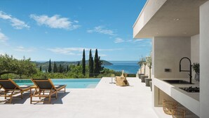 A modern outdoor pool area with lounge chairs, a small table with a drink, and a kitchen area with a sink and countertop. - Lavish Villa O with Panoramic Elefiti Islands View, Six Bedrooms with a Pool (Dubrovnik)