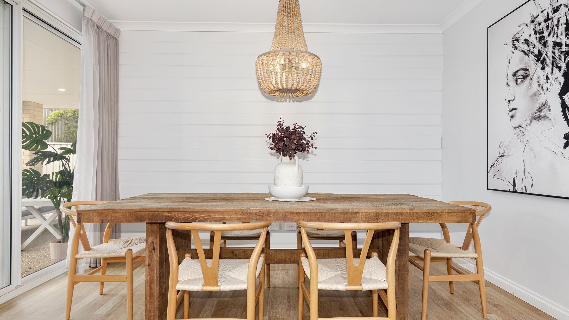 A dining area with a wooden table, six chairs, a white vase with flowers, and a large abstract painting on the wall.