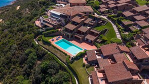 An aerial view of a coastal residential area with terracotta-roofed houses, a central swimming pool, and lush greenery.