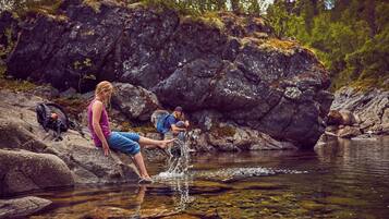 A person sitting on rocks by a clear stream, another person in the background, and a dog nearby.