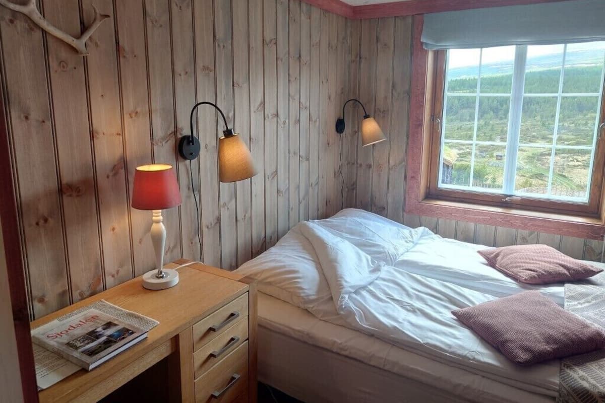 A bedroom with a wooden wall, a bed, a nightstand with a lamp, and a window with a view.