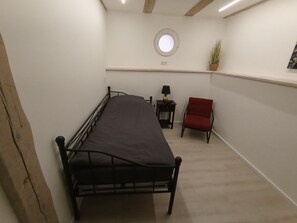 A single bed with a headboard, a small table with a lamp, a red chair, and a potted plant on a white wall. - Alter Speicher (Schönberg)