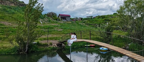 A wooden bridge over a calm body of water, surrounded by lush greenery and a distant building.