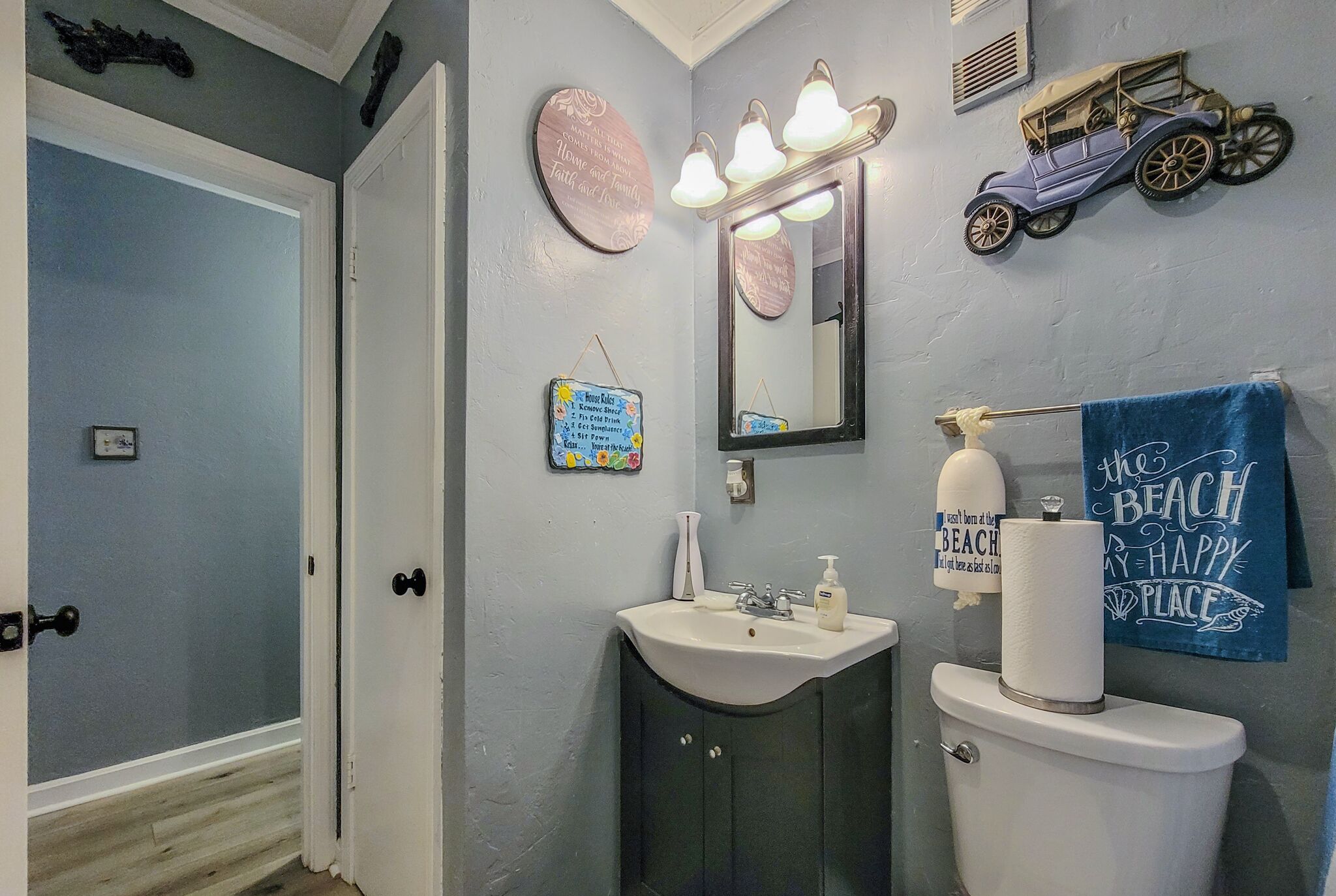 Un baño con lavamanos, inodoro y espejo. Hay elementos decorativos en la pared, incluyendo un coche y un letrero de playa.