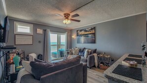 A living room with a grey sofa, a coffee table, a TV, and a ceiling fan. There is a sliding glass door leading to a balcony. - Topsail Reef 358: Dream2B - Oceanfront Getaway! (North Topsail Beach)