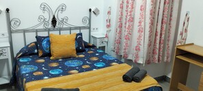 A bedroom with a bed featuring a space-themed bedspread, a wooden headboard, and a pair of rolled towels on the bed.