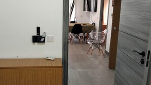 A room with a wooden cabinet, a wall-mounted thermostat, a dining table with chairs, and a door leading to another room.