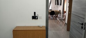 A room with a wooden cabinet, a wall-mounted thermostat, a dining table with chairs, and a door leading to another room.