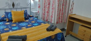 A bedroom with a bed featuring a space-themed bedspread, a bedside table, a wooden desk, and a curtain with a floral pattern.