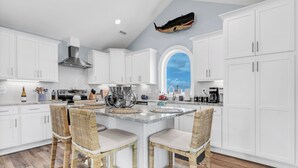 A modern kitchen with white cabinetry, a large island with bar stools, and a unique whale decoration on the ceiling. - Elegant Beach Villa: Pool, Hot Tub, Ocean Views (North Topsail Beach)