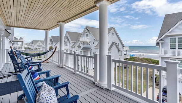 A covered porch with blue chairs and a hammock, overlooking a beach with houses in the background. - Elegant Beach Villa: Pool, Hot Tub, Ocean Views (North Topsail Beach)