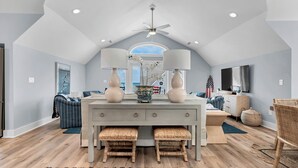 A spacious living room with a high ceiling, a dining area with a white table and two chairs, and a large window offering a view of the sky. - Elegant Beach Villa: Pool, Hot Tub, Ocean Views (North Topsail Beach)