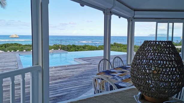 A balcony with a view of the ocean, a pool, and a table with chairs.