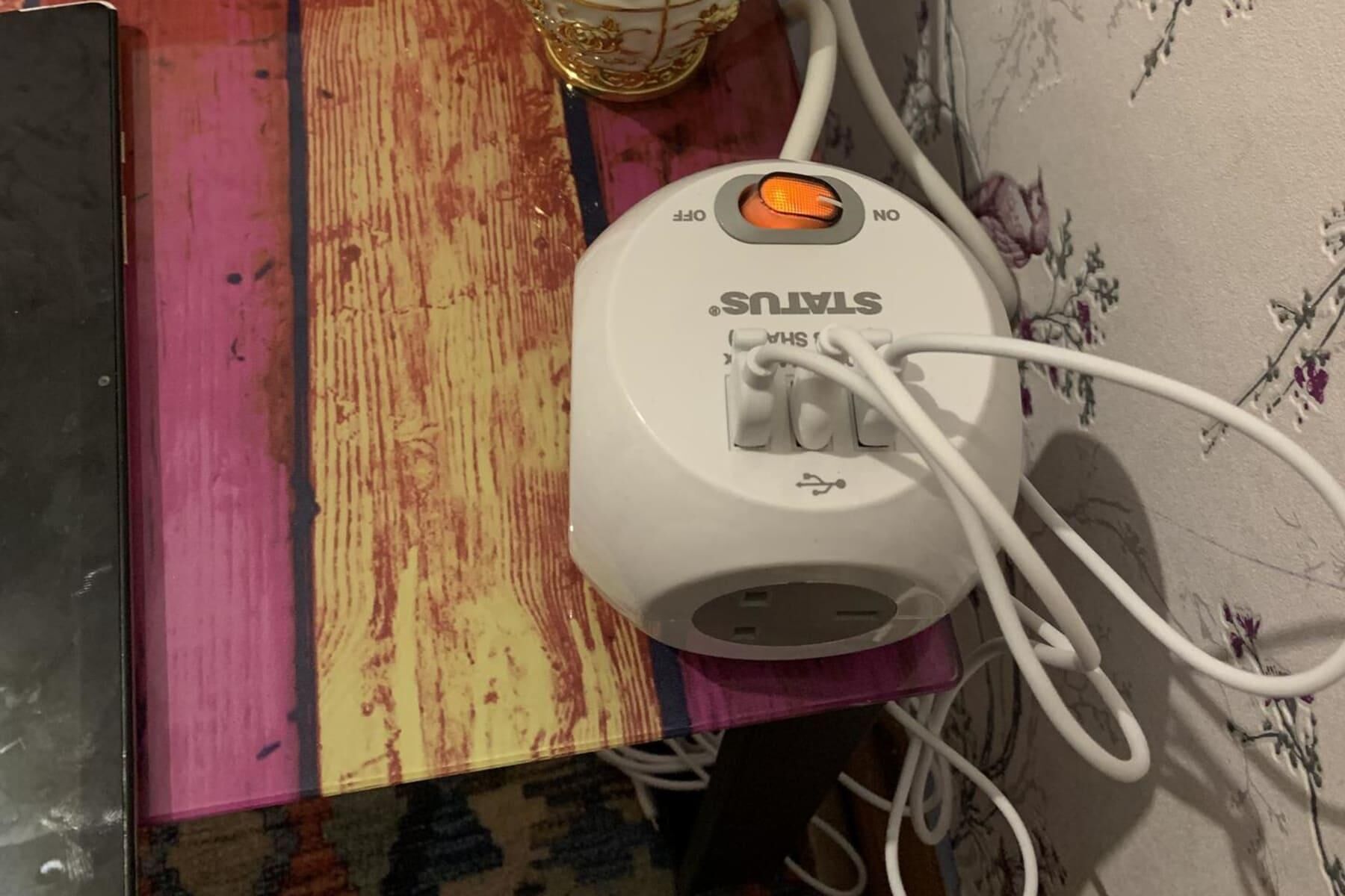 A white power strip with multiple white cables plugged in, placed on a wooden surface with a floral wallpaper background.