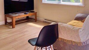 Ett vardagsrum med en platt-TV på ett träställ, en svart stol, en soffa med en gul pläd och ett stort fönster med utsikt över träd.