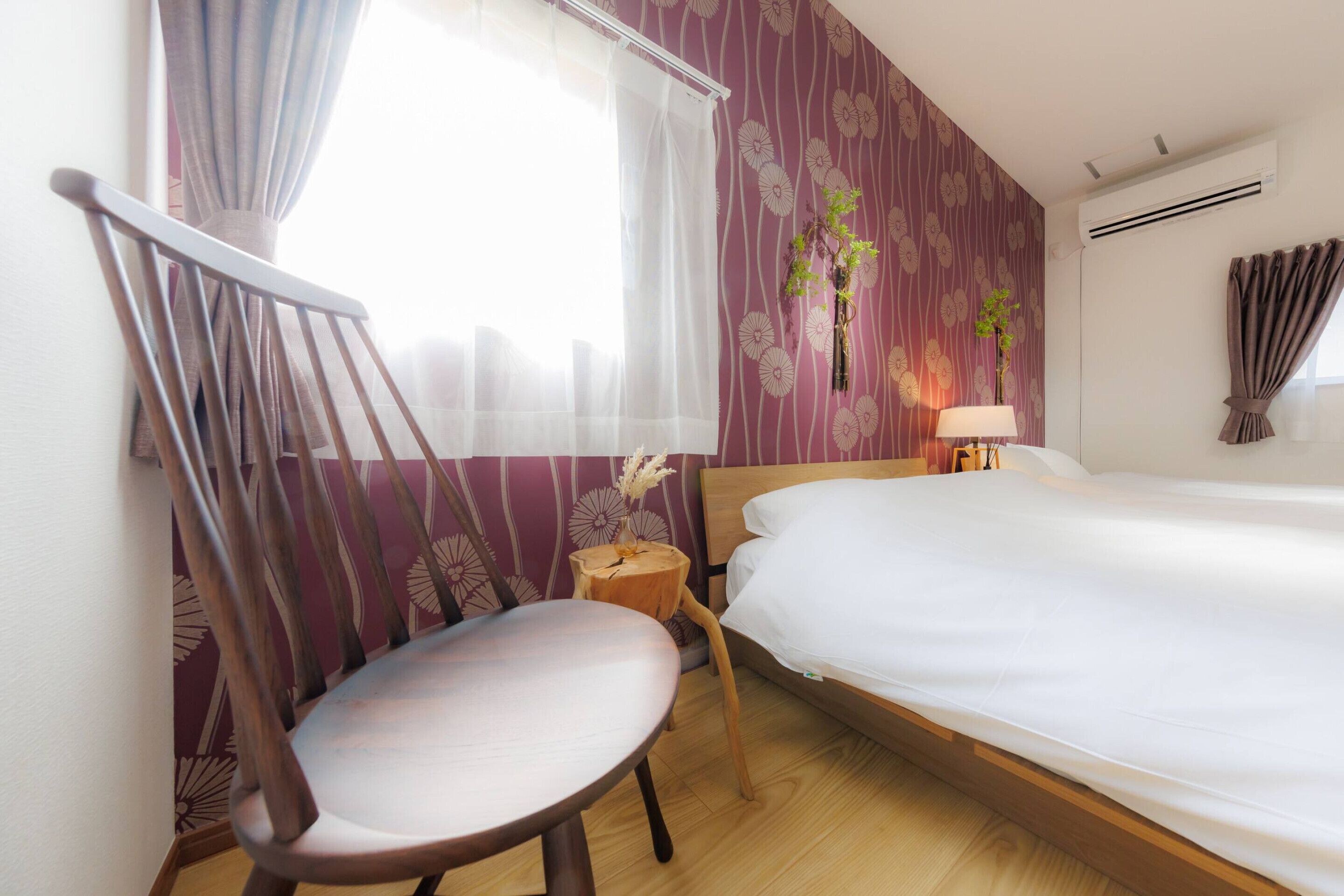 A hotel room with a wooden bed, a chair, a small wooden table, and a window with curtains.