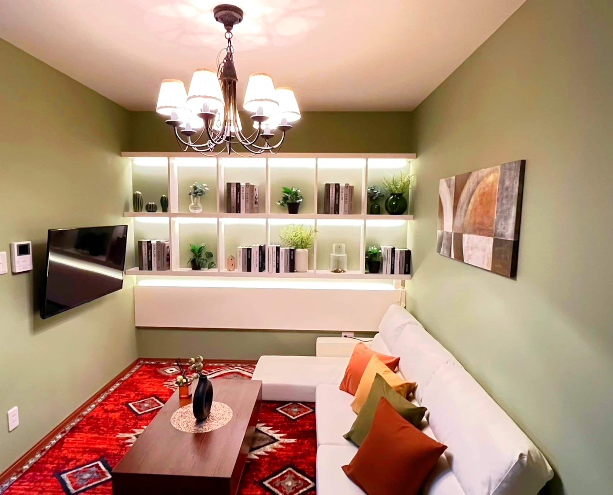 A living room with a flat-screen TV, a white sofa with colorful cushions, a red patterned rug, a wooden coffee table, and a bookshelf with various decorative items.