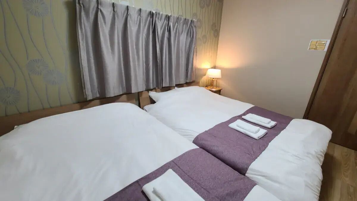 A hotel room with two beds, a nightstand with a lamp, and a curtain.
