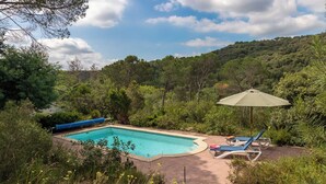 A rectangular swimming pool with a blue tarp on one side, surrounded by lush greenery and a hillside. There are two lounge chairs and a large umbrella. - Charming Home in a Green Valley. El Pigot. Tranquility, Nature & Private Pool! (Riudarenes)