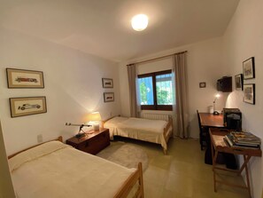 A small, well-lit bedroom with two single beds, a desk with a lamp, a radiator, and framed pictures on the wall.