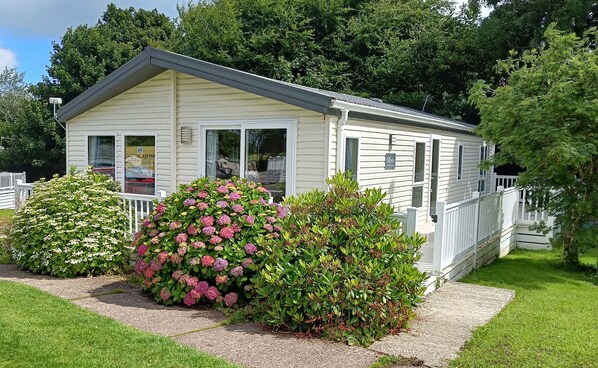 A small, single-story mobile home with a white exterior and a grey roof. It features a front porch with a white railing and a large bush with pink flowers.