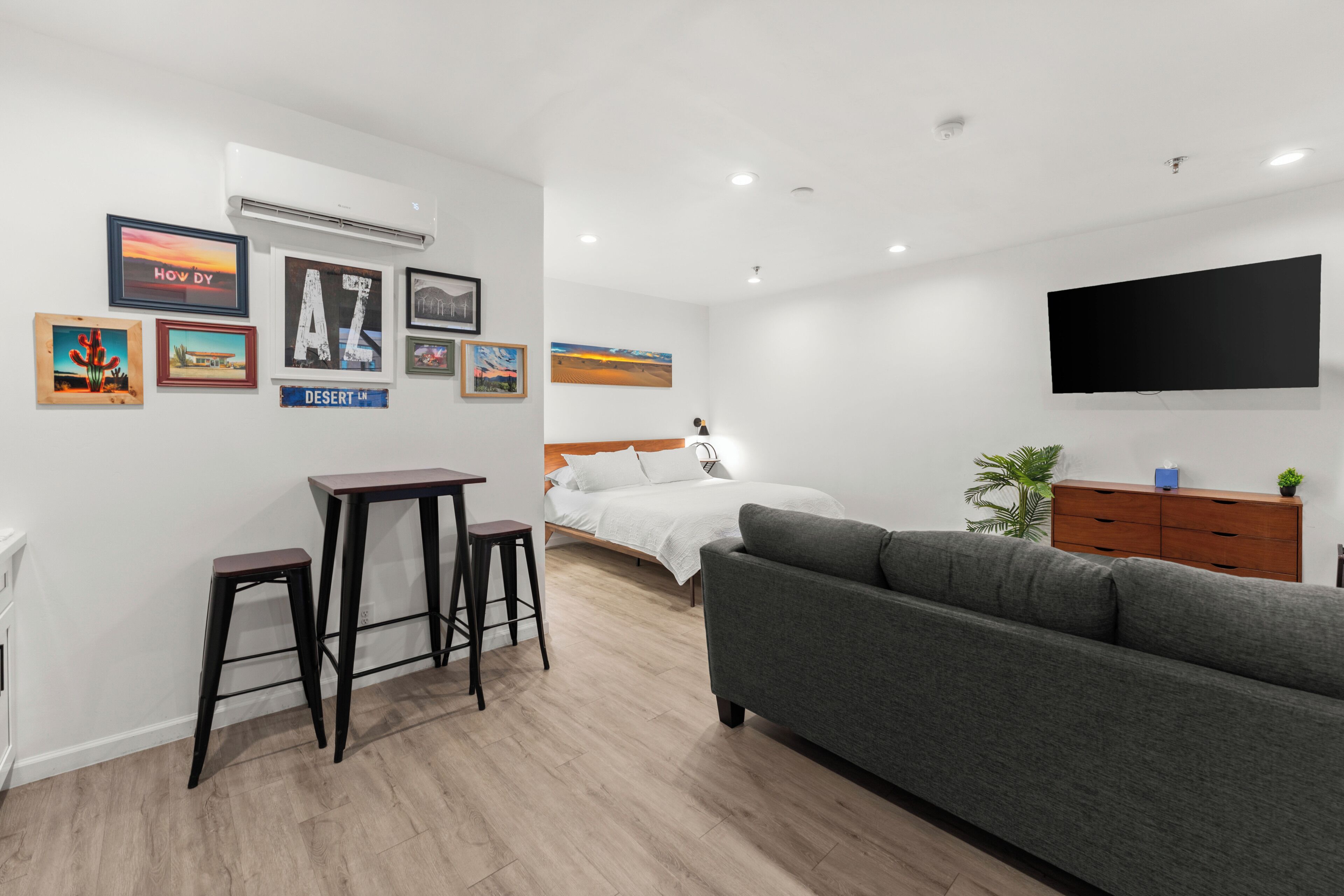 Una habitación de hotel moderna con una cama, un sofá gris, un escritorio con dos taburetes, un televisor de pantalla plana y varias imágenes enmarcadas en la pared.