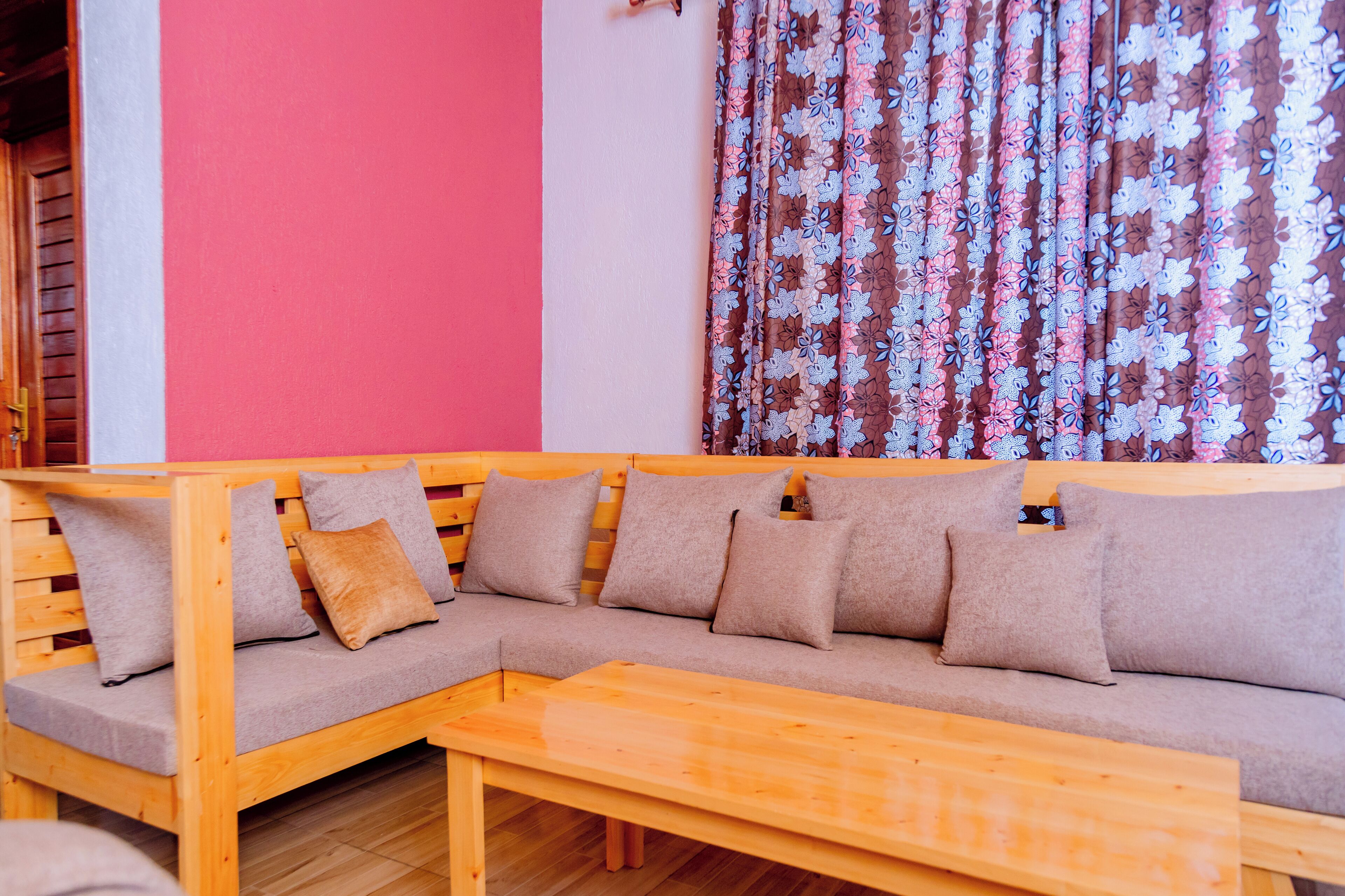 A wooden sectional sofa with cushions, a wooden table, and a red wall with a floral curtain.