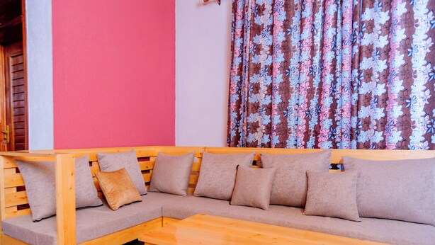 A wooden sectional sofa with cushions, a wooden table, and a red wall with a floral curtain. - Future4kids Kinigi Guest House By Isange Paradise Resort (Musanze)