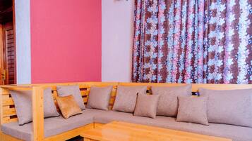 A wooden sectional sofa with cushions, a wooden table, and a red wall with a floral curtain.