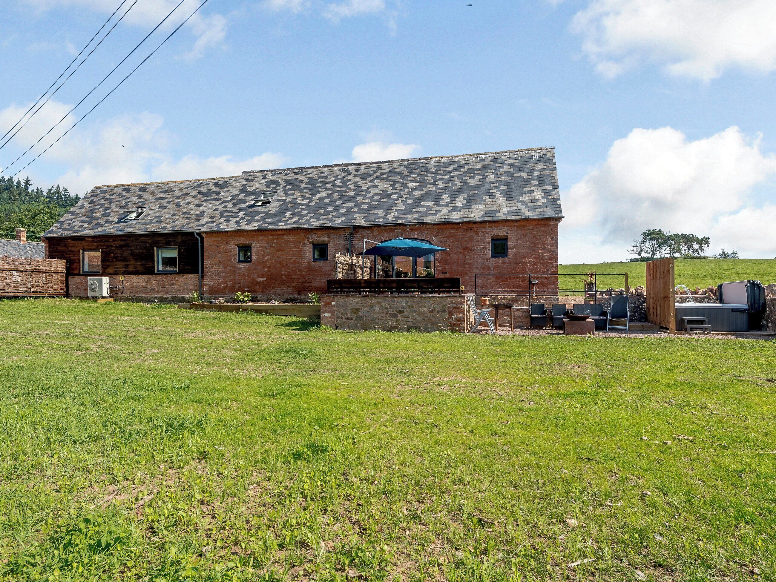 Un bâtiment rustique en briques, doté d’un toit en pente, d’un patio avec des sièges extérieurs et offrant une vue sur un champ verdoyant.