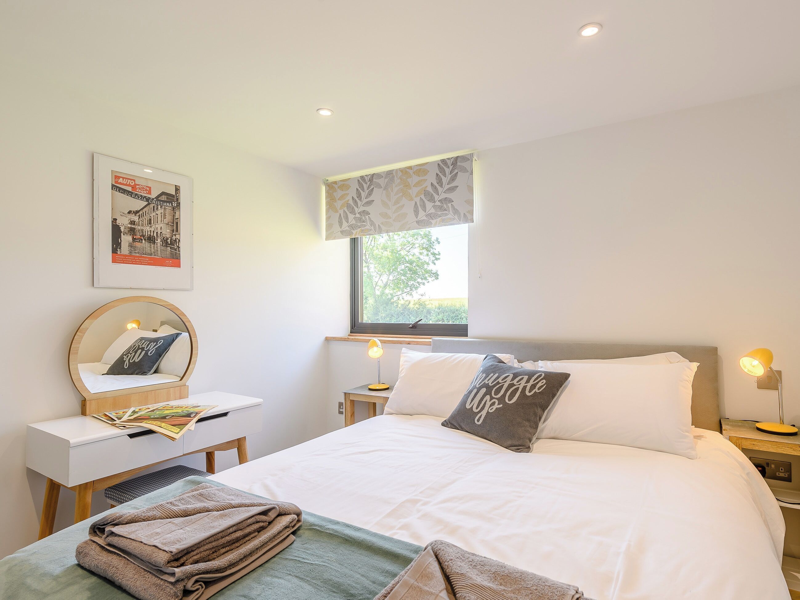 A neatly arranged bedroom with a bed, bedside tables, a desk, a chair, a window with a view, and a poster on the wall.